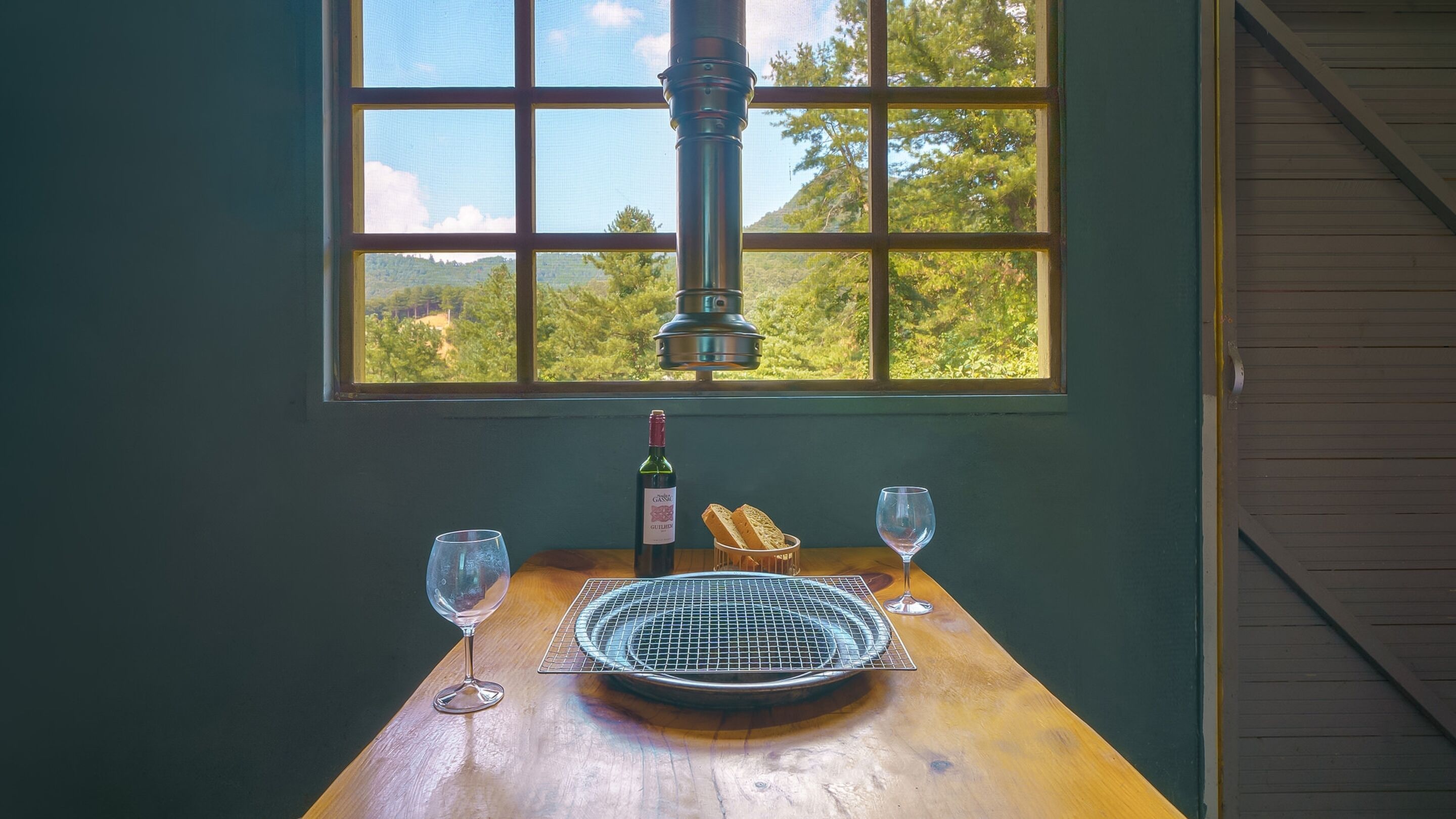 A table with a bottle of wine, two glasses, and a bread basket in front of a window with a view of trees.
