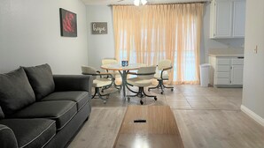 Een moderne woonkamer met een zwarte bank, een eettafel met stoelen, een plafondventilator en een keuken met kastjes.