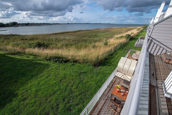 Une vue depuis un balcon donnant sur une zone herbeuse, avec un plan d’eau et une côte lointaine.