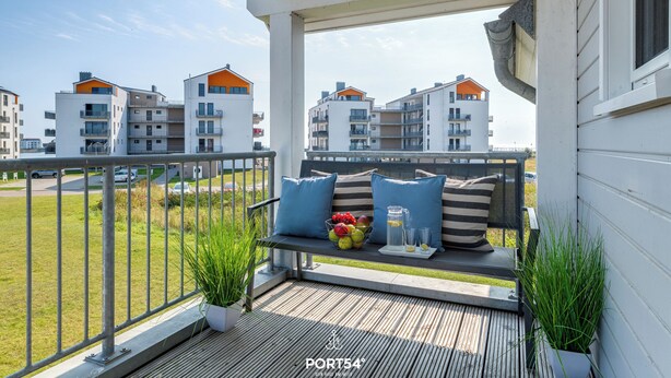 Un balcon doté d’un plancher en bois, d’une rambarde en métal et d’un banc agrémenté de coussins et d’un panier de fruits.