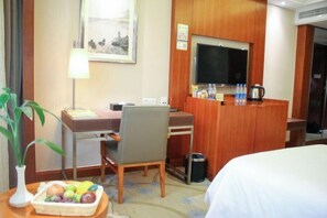 A hotel room with a bed, a desk with a chair, a television, a small table with a fruit basket, and a trash bin. - Joyful Hotel (Yueqing)