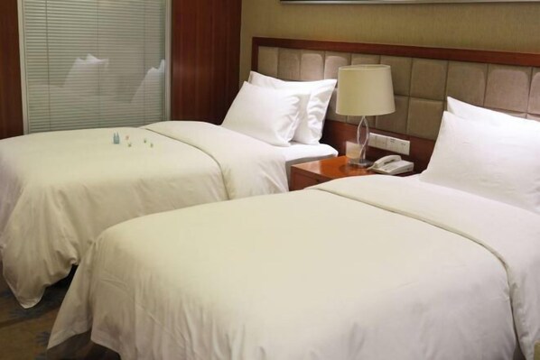 Two beds with white linens, a wooden headboard, a bedside table with a lamp, and a window with blinds. - Joyful Hotel (Yueqing)