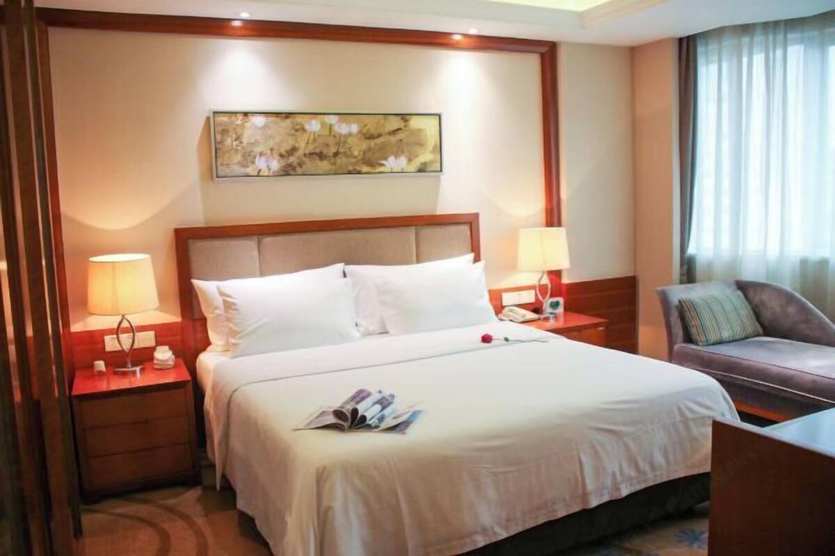 A hotel room with a large bed, two bedside tables with lamps, a small table with a telephone, a sofa with a cushion, and a painting on the wall.