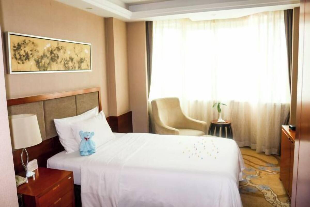 A hotel room with a bed, a chair, a desk, a lamp, a painting, and a window with curtains.
