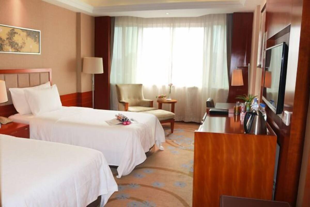 A hotel room with two beds, a desk with a TV, a chair, a lamp, and a window with curtains.