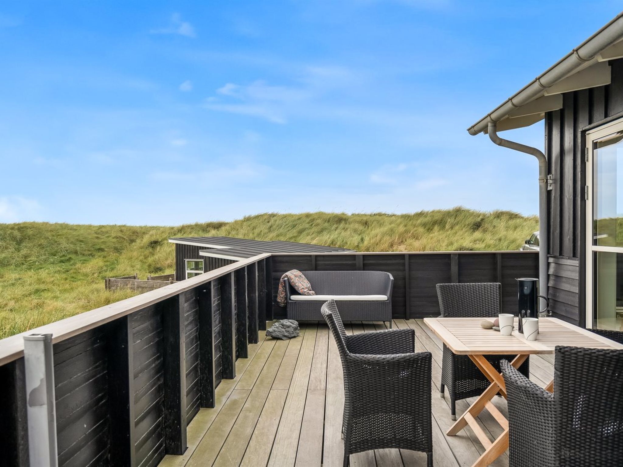 Een terras met buitenmeubilair, een houten tafel en stoelen, met uitzicht op een duinlandschap.