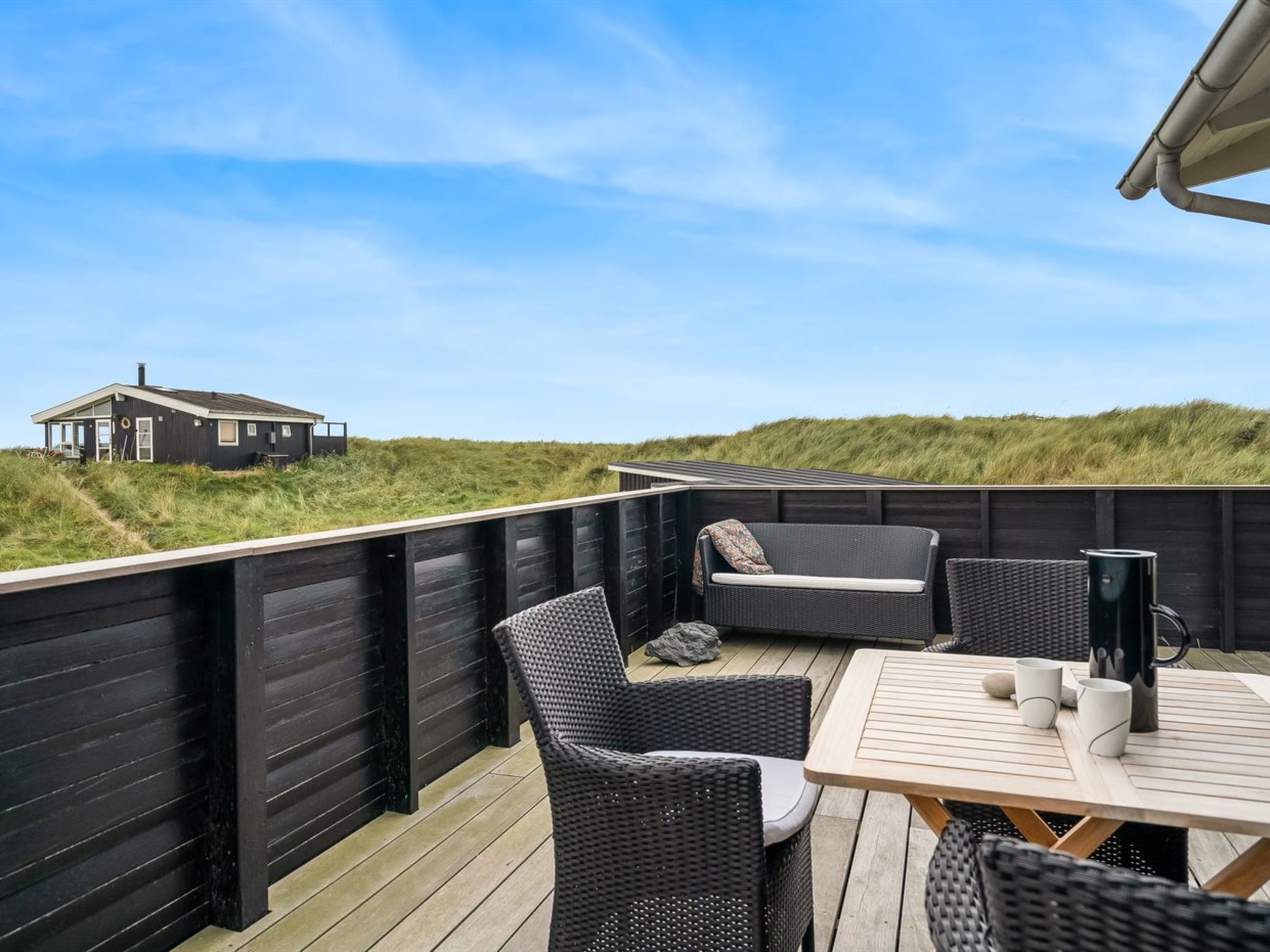 A wooden deck with wicker furniture and a table, overlooking a grassy landscape and a house.