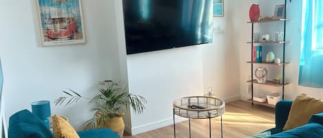 A living room with teal sofas, a flat-screen TV, a bookshelf, and a potted plant.