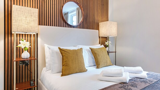 Hotelkamer met een houten paneelwand, een ronde spiegel, een bed met wit beddengoed, twee kussens, een nachtkastje met een lamp en een vaas met bloemen.