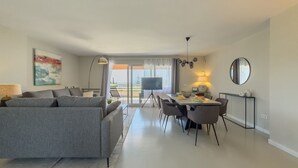 Una sala de estar moderna con un sofá gris, una mesa de comedor con sillas, un televisor de pantalla plana y un ventanal con cortinas.