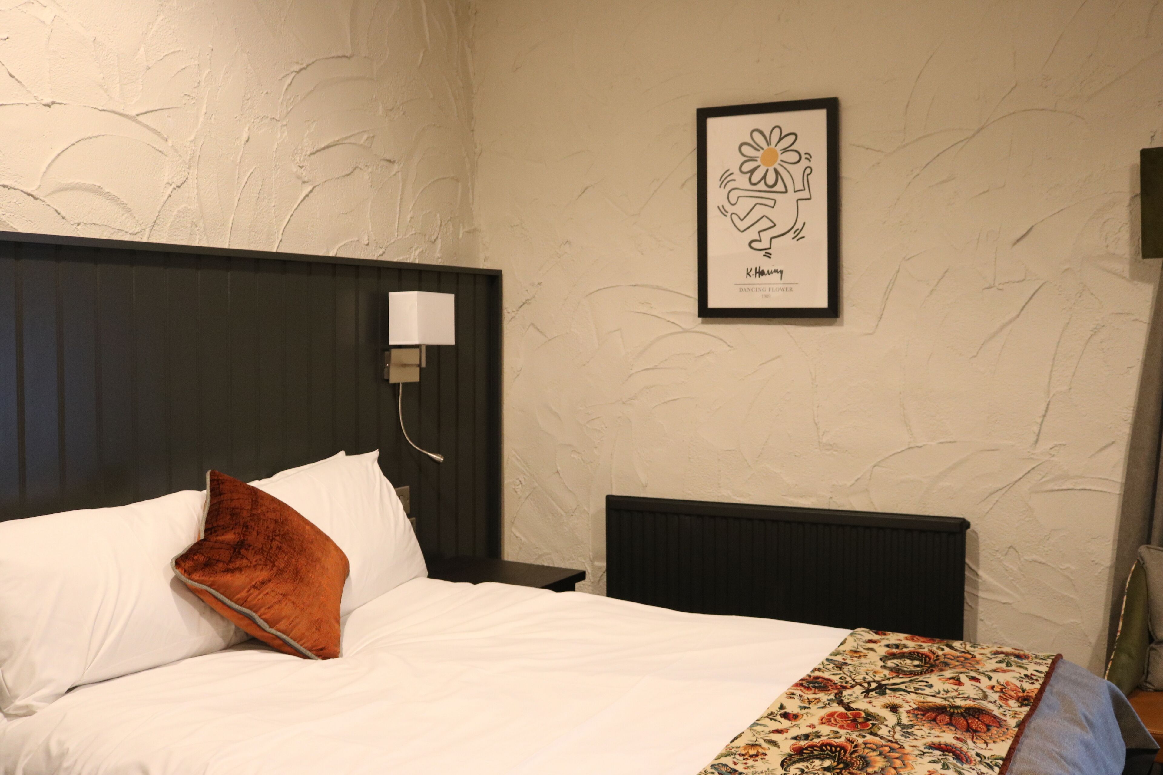A neatly made bed with white linens and a brown pillow, a framed artwork on the wall, and a nightstand with a lamp.