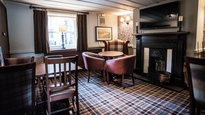 A cozy room with a checkered floor, a fireplace, a television mounted above it, and a dining area with chairs and a table. - Black Dog Inn (Bolton)