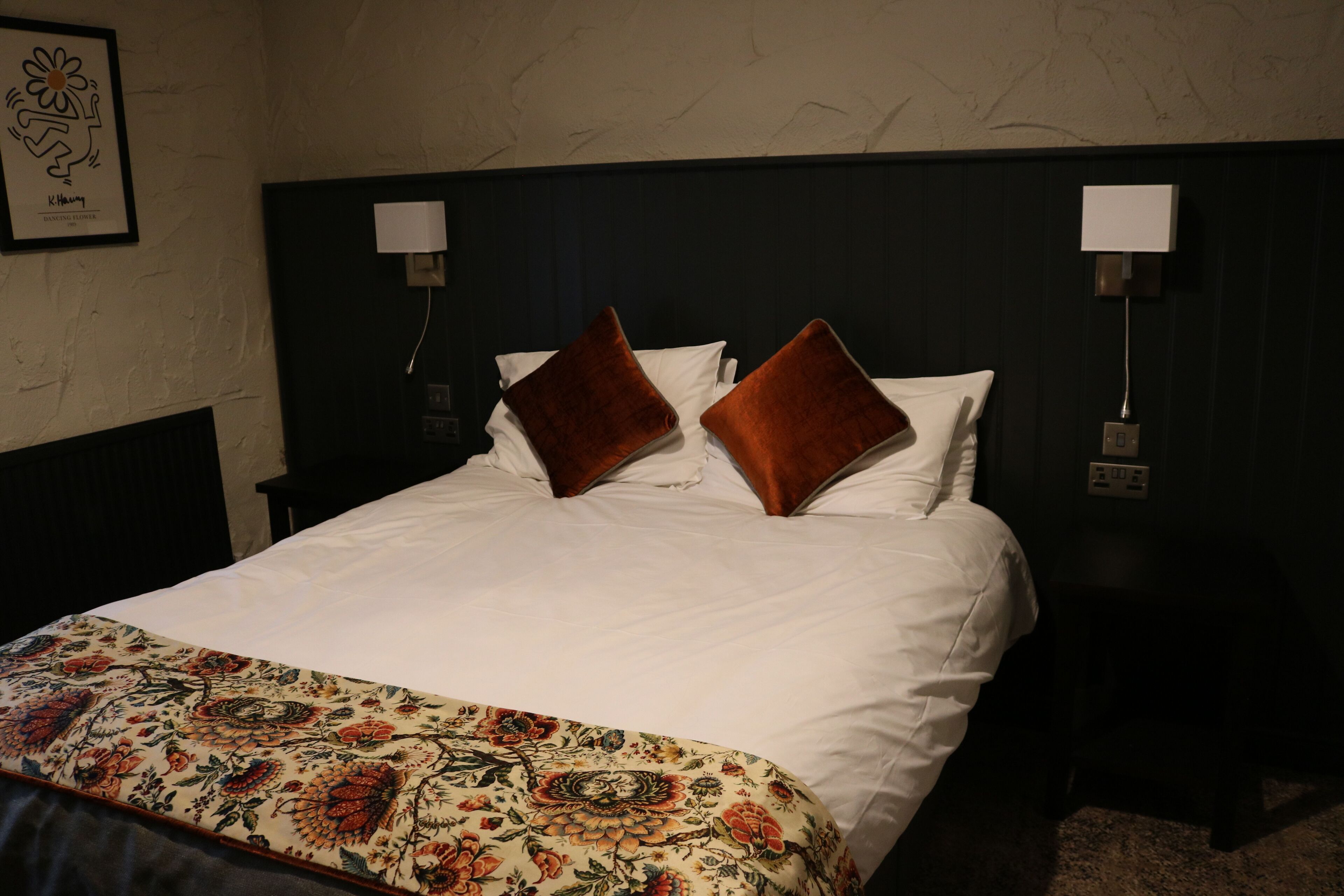 Un lit bien fait, recouvert d’une couvre-lit à motifs floraux, avec deux oreillers marron et une tête de lit agrémentée de lampes fixées au mur.