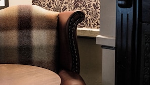 A corner of a room with a plaid-patterned armchair, a round wooden table, and a patterned wallpaper. - Black Dog Inn (Bolton)