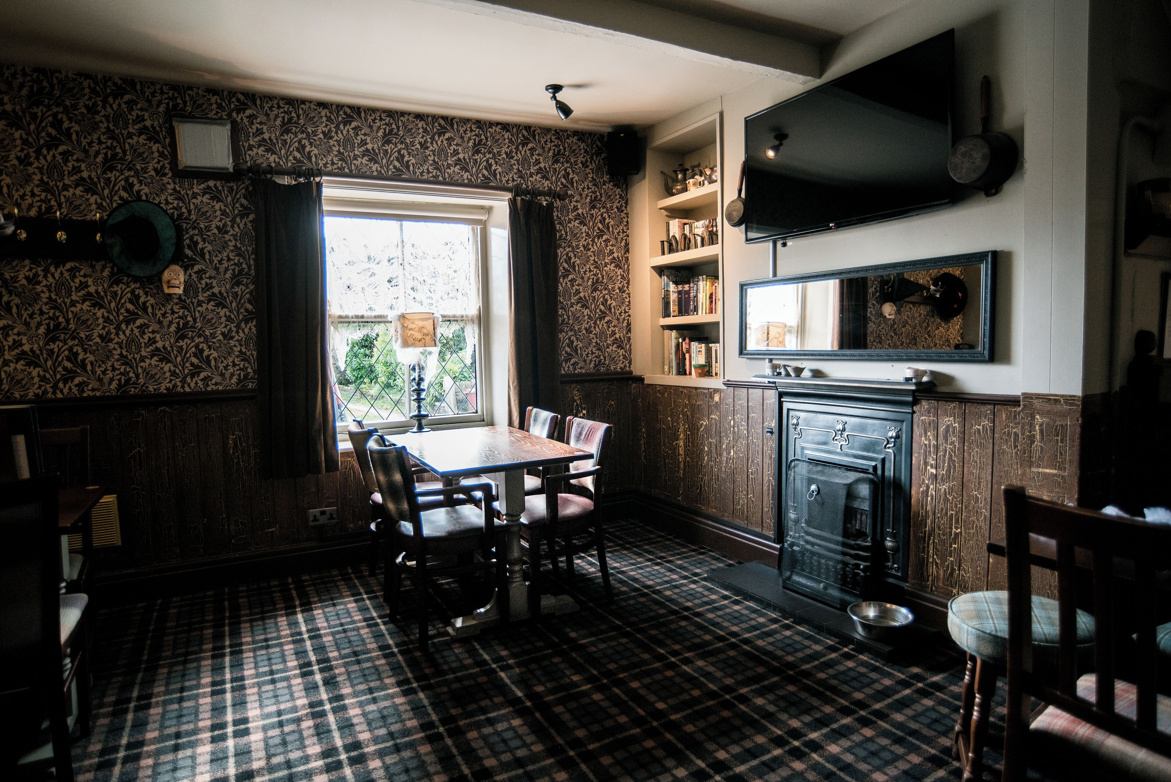 A dining area with a checkered floor, a wooden table, and chairs. There is a fireplace with a mirror above it and a television mounted on the wall.