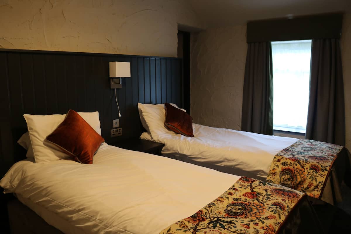 A hotel room with two beds, each with white bedding and a red pillow. There is a nightstand with a lamp between the beds. The room has a window with curtains, and the walls are textured.