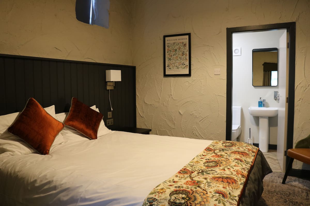 A hotel room with a bed, a nightstand, a wall-mounted lamp, a framed picture, a bathroom visible through an open door, and a chair.