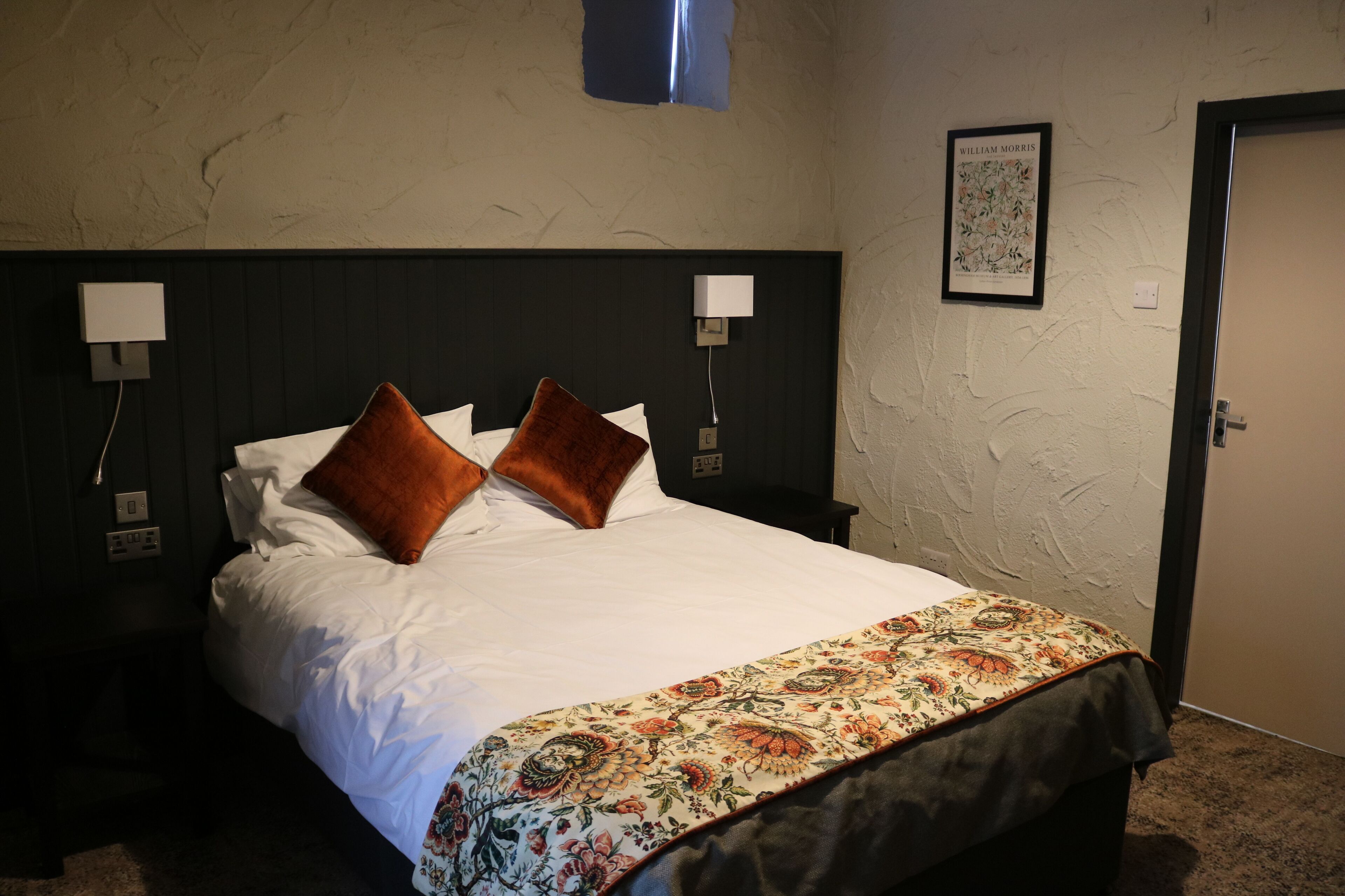 A neatly made bed with a floral bedspread and two brown pillows, a dark headboard, two wall-mounted night lights, and a framed picture on the wall.