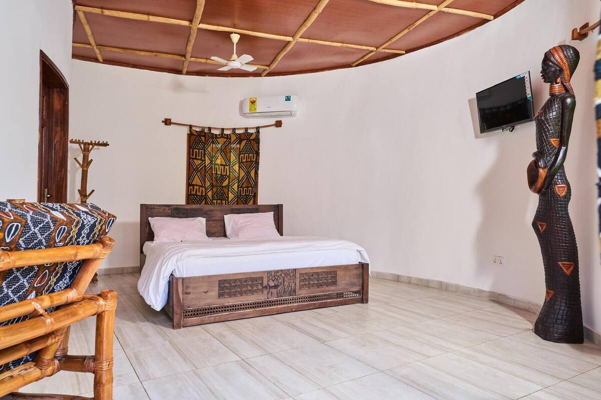 A room with a bed, a wooden chair, a ceiling fan, an air conditioning unit, a wall-mounted TV, and a decorative sculpture.