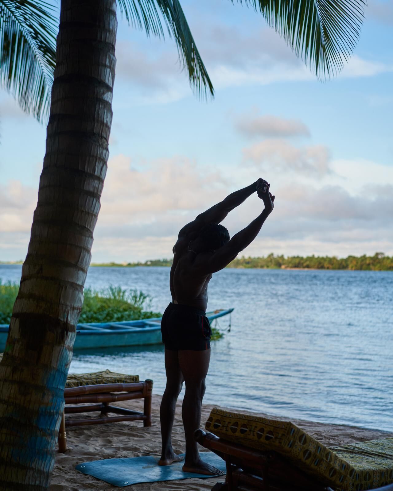 A person stretching on a mat by a body of water with boats and a palm tree in the background.
