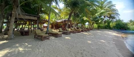Una playa con sillones de madera y una zona de estar cubierta bajo palmeras.