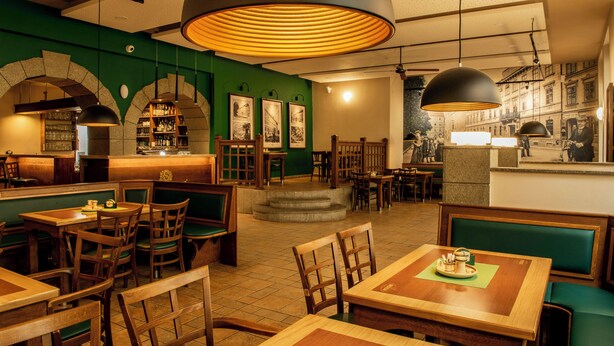 A restaurant interior with wooden tables and booths, green upholstered seating, and a bar area with shelves of bottles. - Beseda Accomodation (Ceské Budejovice)
