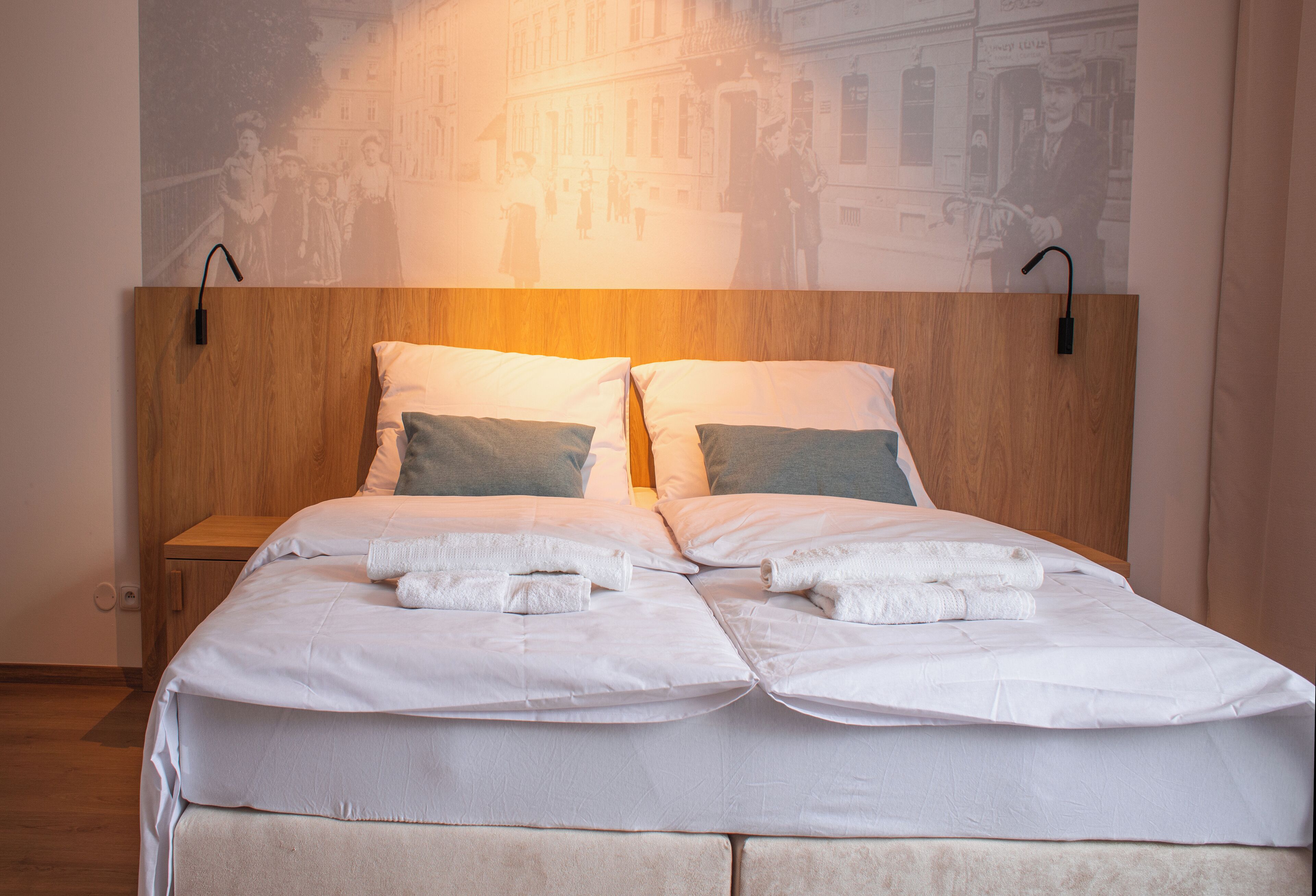 A neatly made bed with white linens and two pillows, a wooden headboard, and a wall-mounted light fixture.