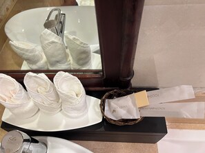 Un lavamanos de baño con un lavabo blanco, un espejo y una bandeja con toallas dobladas. Hay dos toallas blancas sobre la bandeja y una cesta pequeña con pañuelos de papel y una tarjeta.