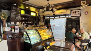 A coffee shop interior with a menu board, a glass display case with pastries, and a seating area with tables and chairs.
