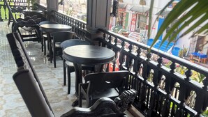 A covered outdoor seating area with black metal chairs and tables, a decorative railing, and a view of the street with buildings and shops.