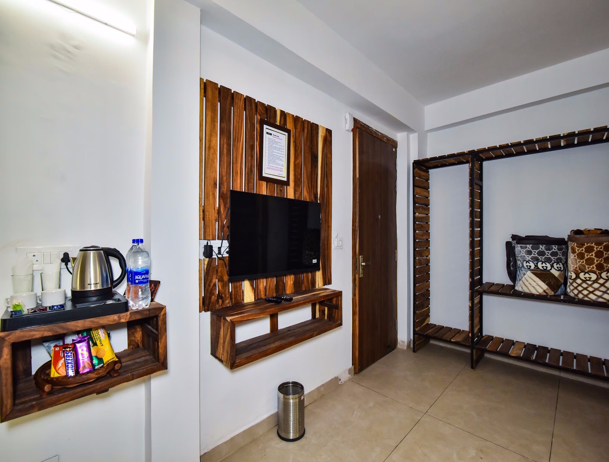 A room with a mounted TV, wooden shelves, a coffee maker, and a trash bin.