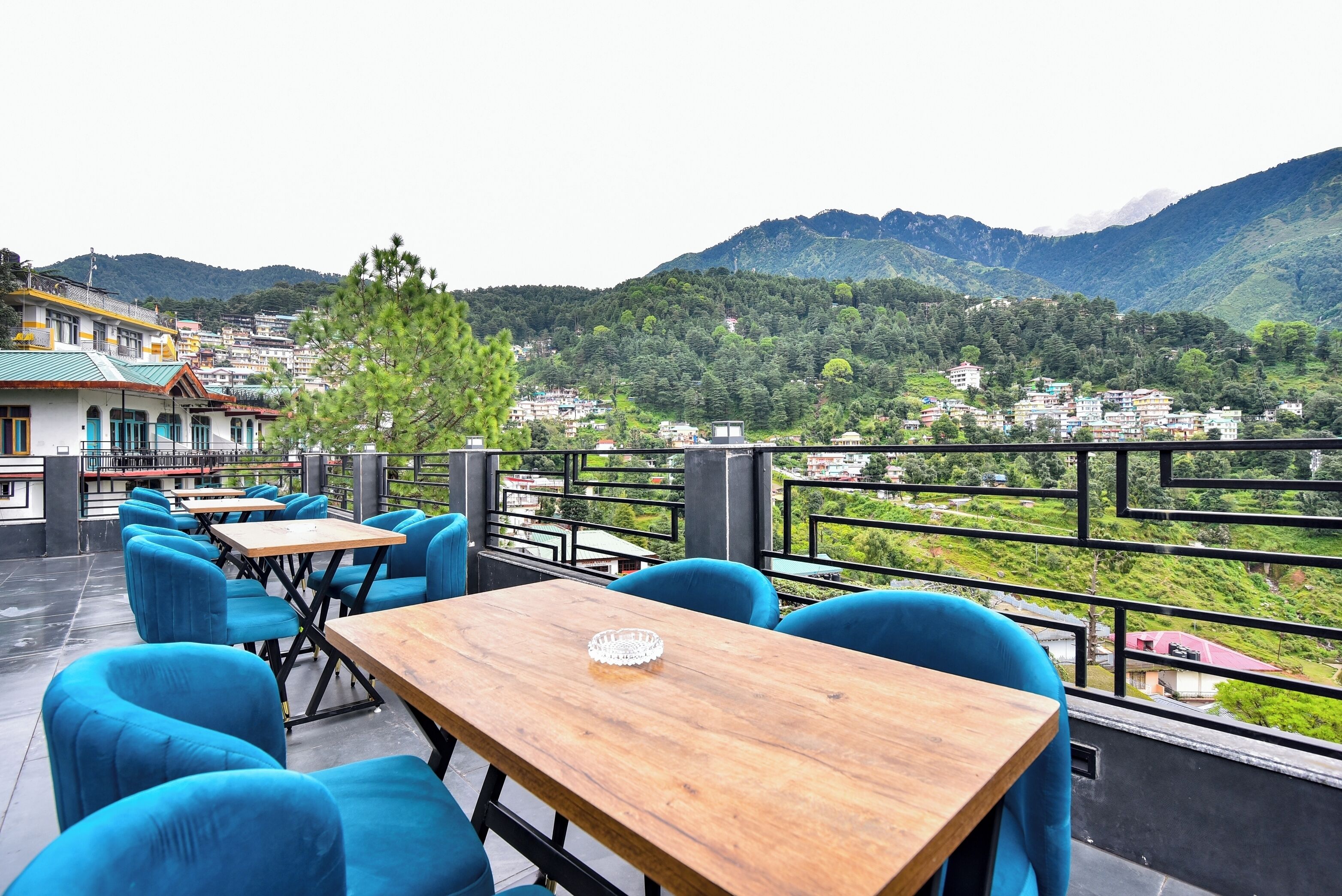 Una terraza con mesas de madera y sillas azules, con vistas a un paisaje montañoso y edificios a lo lejos.