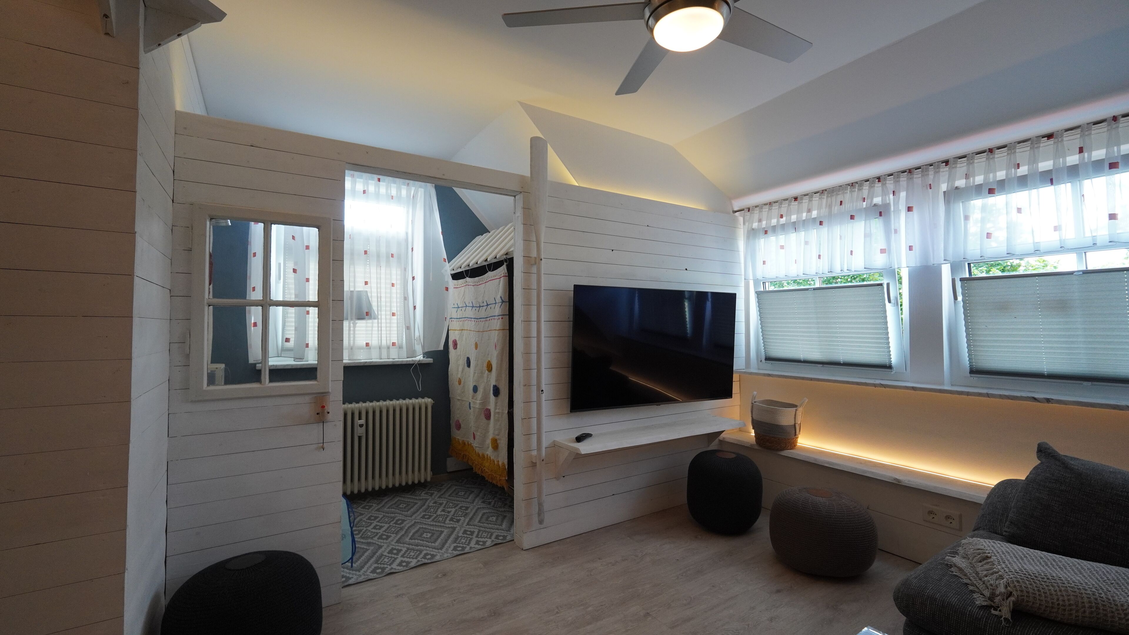 A modern living room with a flat-screen TV mounted on the wall, a ceiling fan, and a large window with blinds.
