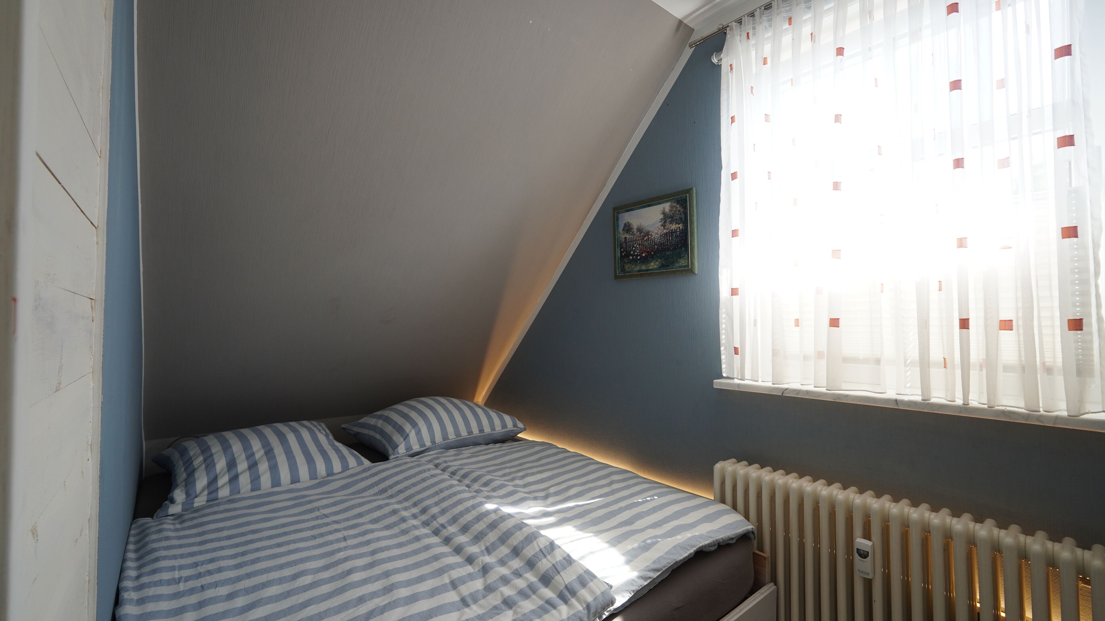 Ein kleiner, dreieckiger Raum mit einem Bett, einem Heizkörper und einem Fenster mit durchsichtigen Vorhängen.