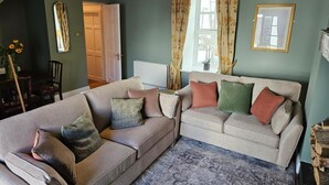 Ein Wohnzimmer mit einer Couch, einem Zweisitzer-Sofa, einem Spiegel, einem Fenster mit Vorhängen und einer Tür.