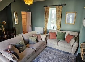 Una sala de estar con un sofá y un sillón, un espejo, una ventana con cortinas y una puerta.