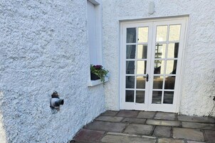 Un edificio blanco con puerta y ventana blancas, escalones de piedra y un pequeño ventanal con flores.