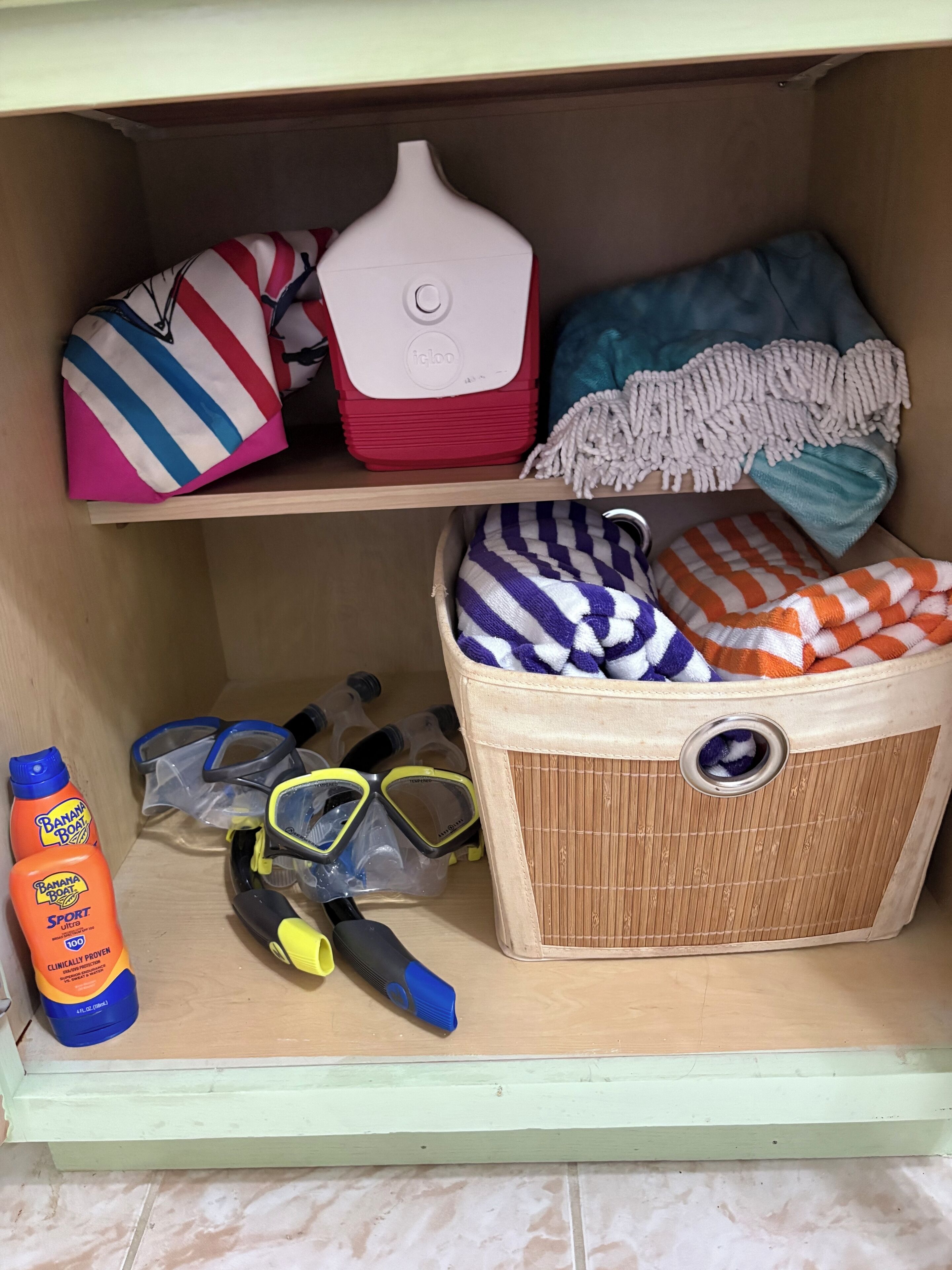 A storage area with a red and white container, a blue and white striped bag, a blue and white checkered bag, a blue and white striped towel, a blue and white towel, a yellow and black snorkel, a pair of black and yellow goggles, and two bottles of Banana Boat Sport.