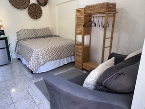 A bedroom with a bed, a wooden wardrobe with hanging clothes, a grey sofa, and a small desk with a lamp. - Cozy Studio In Center of Island • Close to Health City, Beaches and  Restaurants (Bodden Town)