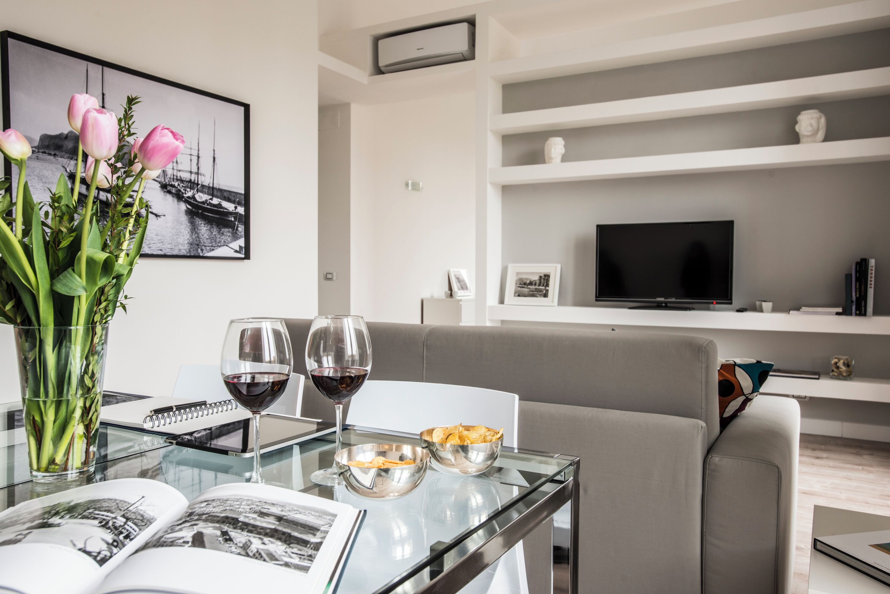 A modern living room with a glass coffee table, a book, a glass of red wine, and a vase of pink tulips.