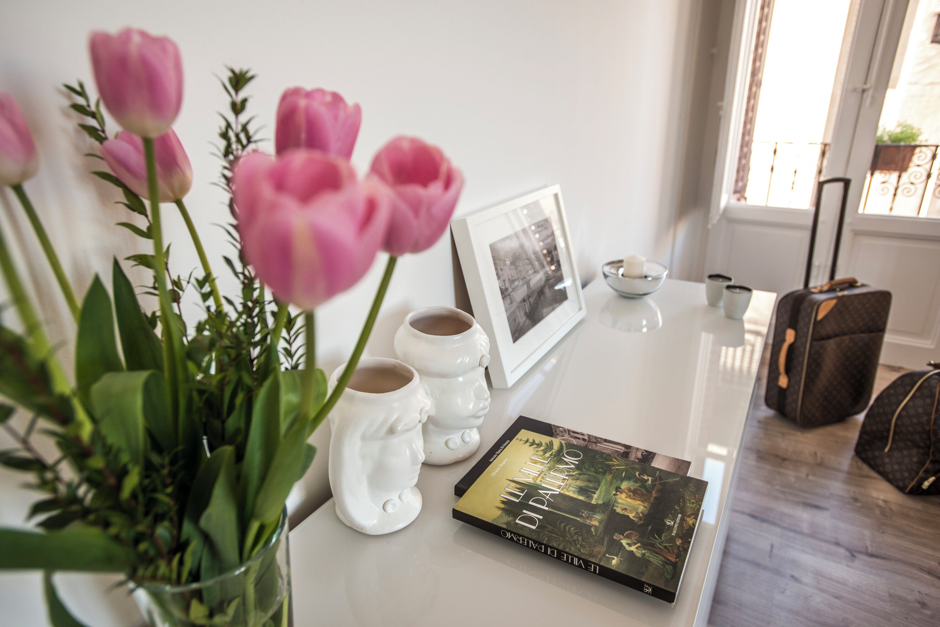 Uma mesa com um livro, uma fotografia emoldurada e um vaso de tulipas cor-de-rosa.
