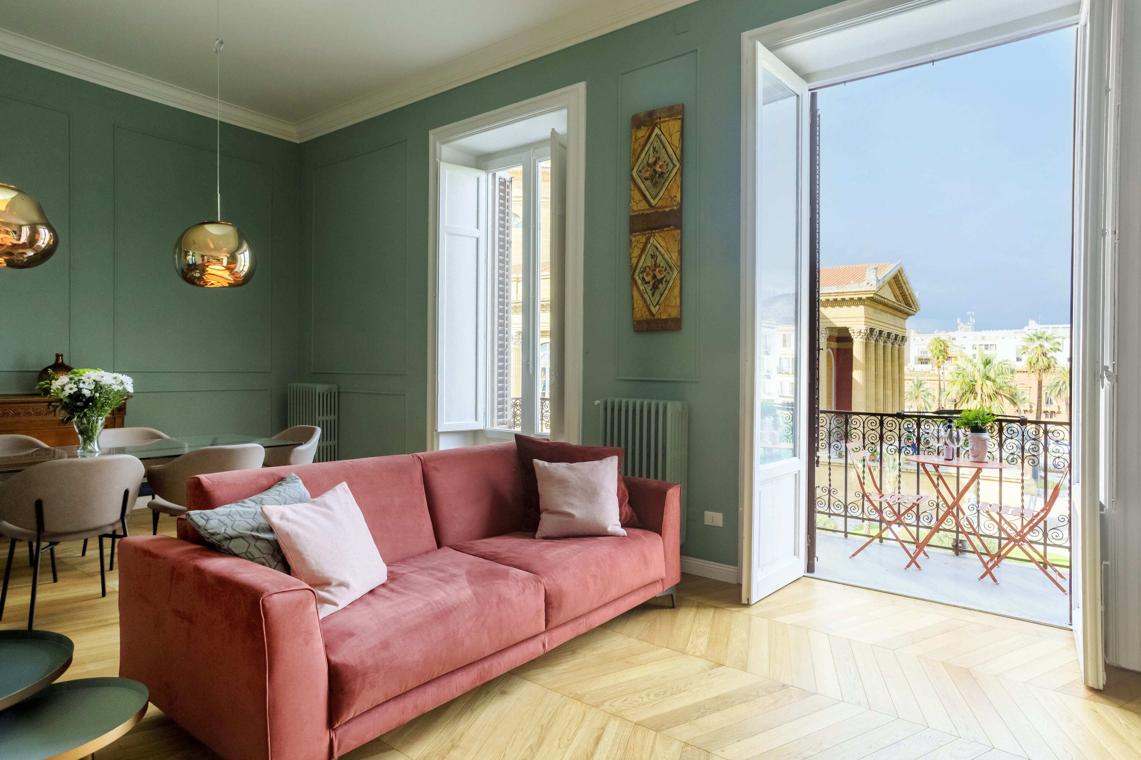 A living room with a pink sofa, a dining table with chairs, and a balcony with a view of buildings and palm trees.