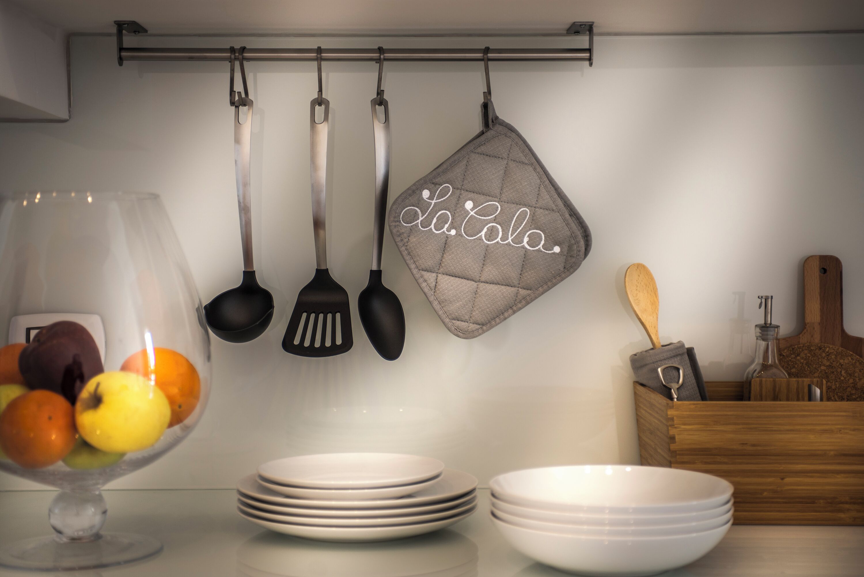 A kitchen with hanging utensils, a fruit bowl, stacked plates, and a wooden box with kitchen tools.