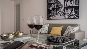 A modern living room with a glass coffee table, two wine glasses, bowls, and a magazine. A sofa with patterned cushions and a cityscape painting on the wall are visible.