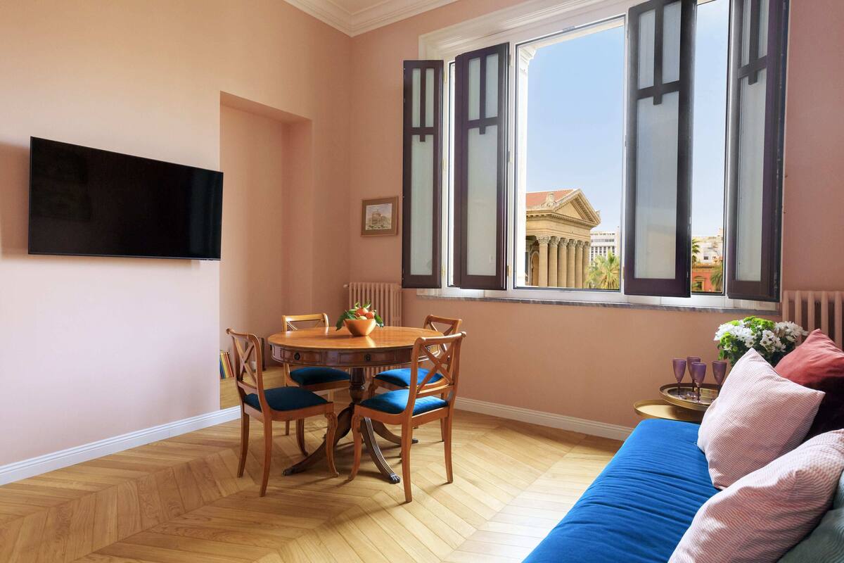 Une pièce comprenant une table à manger, des chaises, un canapé bleu et une fenêtre dont les volets sont ouverts.