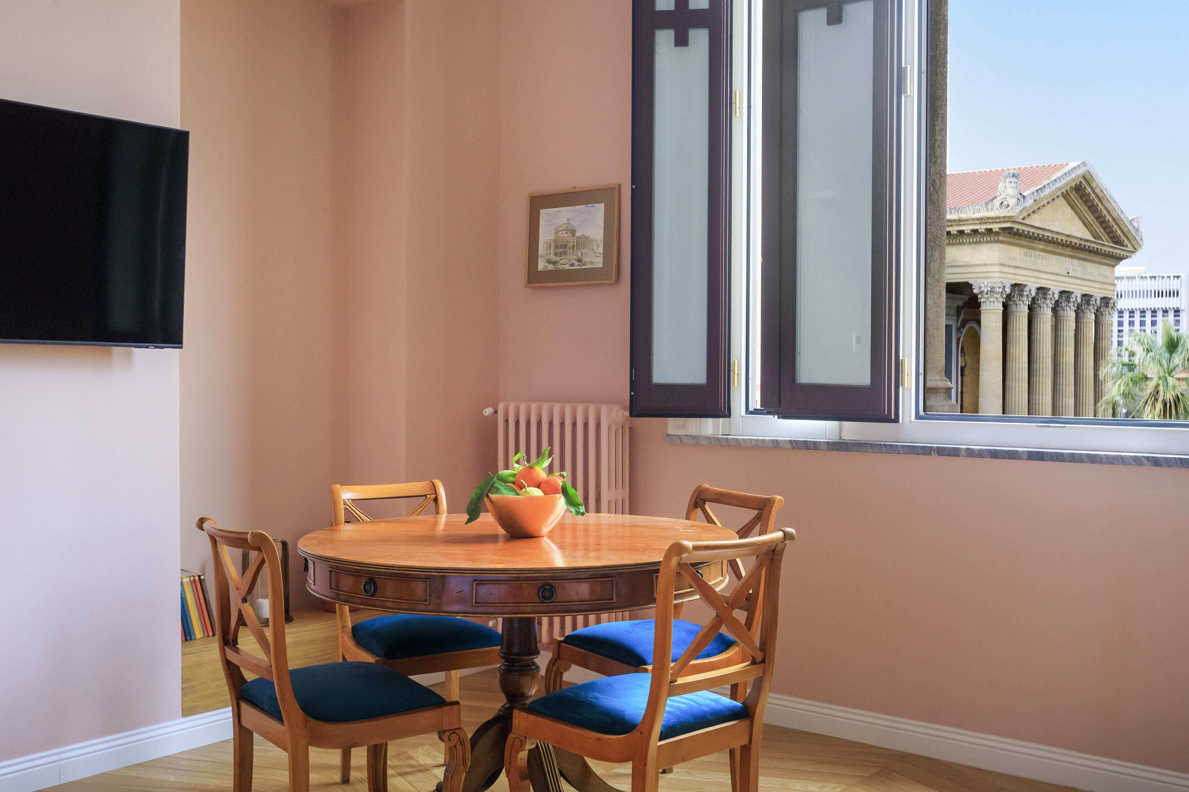 Un comedor con una mesa redonda de madera, sillas con cojines azules, un frutero y un cuadro en la pared.