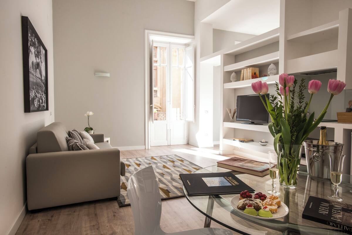 Un salon moderne avec un canapé, une table basse en verre, un vase de fleurs et une bibliothèque.