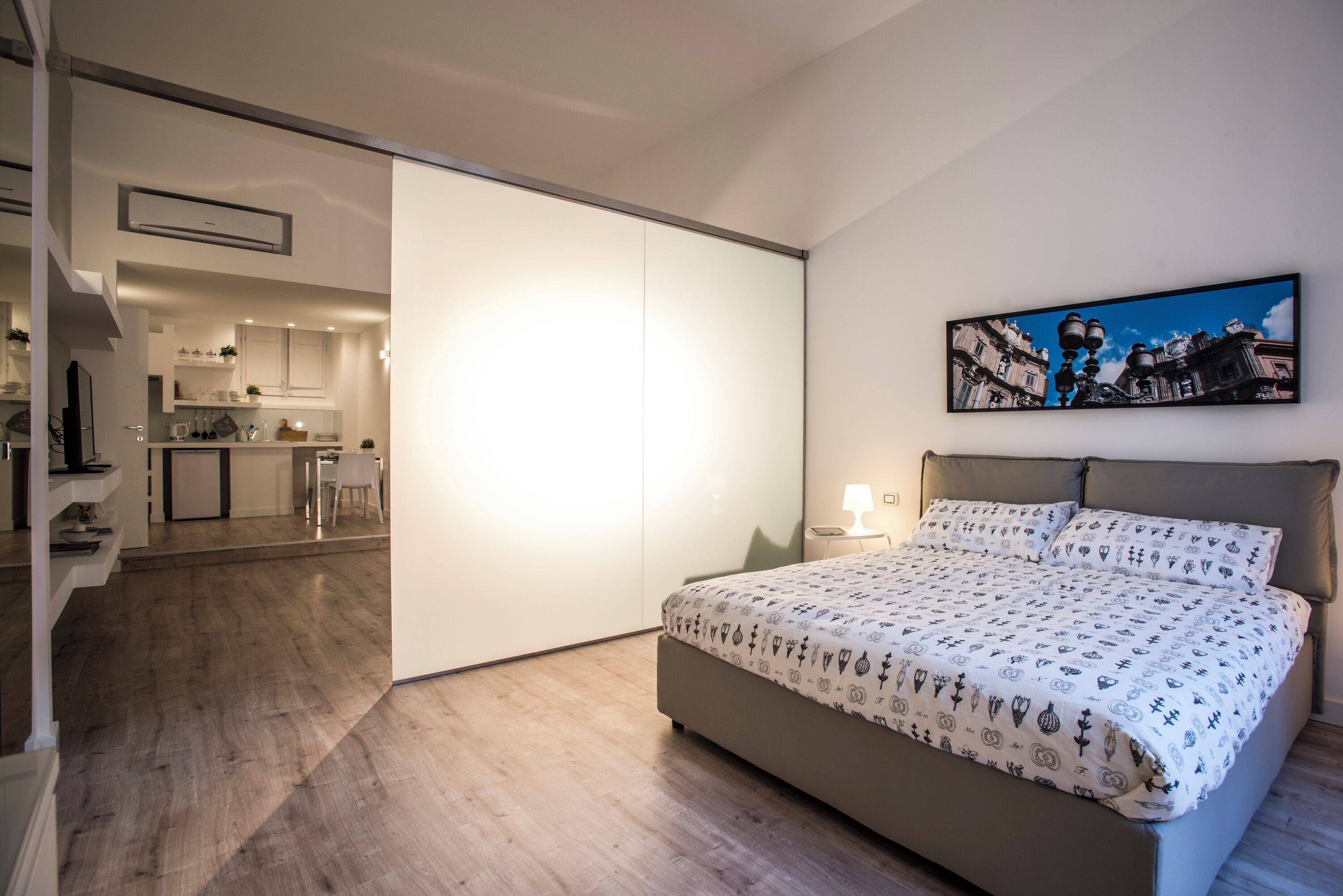A bedroom with a bed, a nightstand, a painting, and a view of a kitchen and dining area.