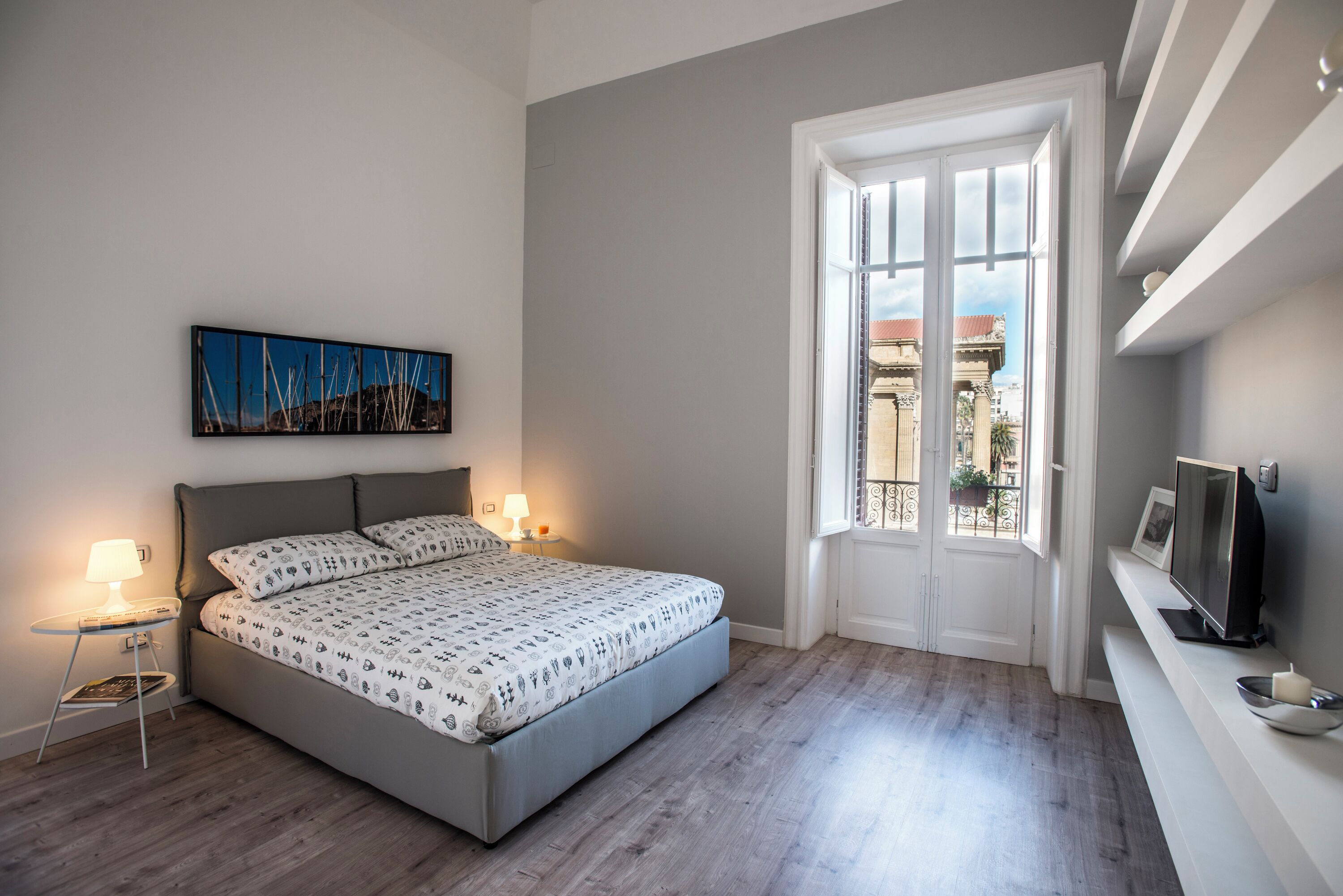 A bedroom with a bed, bedside lamps, a TV, and a window with a view of buildings.