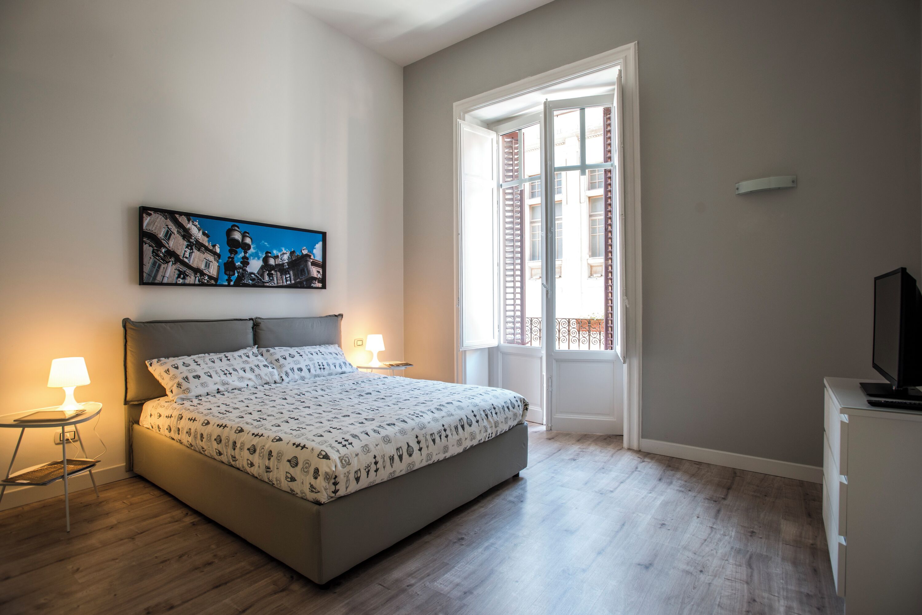 A bedroom with a bed, bedside lamps, a television, a window with shutters, and a framed picture on the wall.